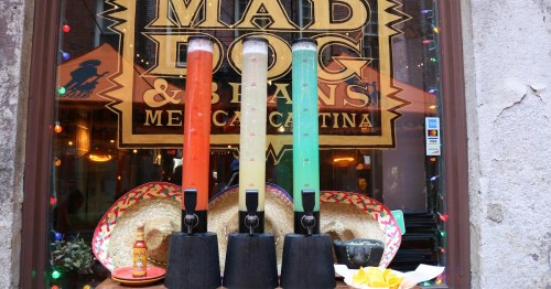 Three colorful drink dispensers in front of a restaurant window, with sombreros and snacks on display.