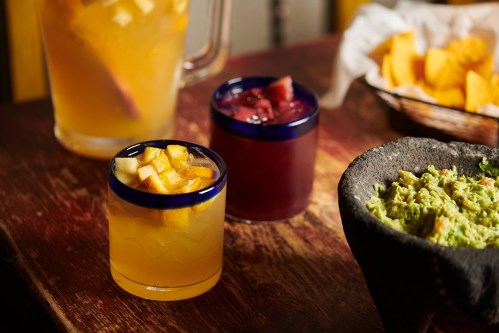 Two colorful cocktails with fruit garnish sit on a wooden table, alongside a bowl of guacamole and a basket of chips.