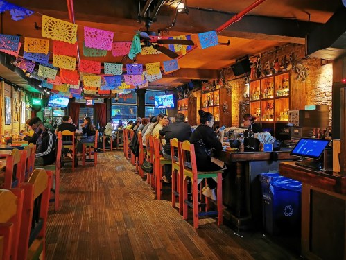 A lively restaurant interior with colorful papel picado decorations, patrons at the bar, and a warm, inviting atmosphere.
