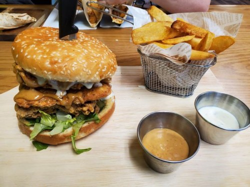 A large sesame seed burger stacked with fried chicken, lettuce, and sauces, served with a side of golden fries.