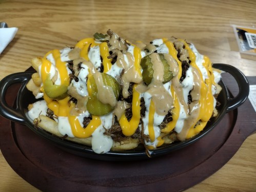 A loaded plate of fries topped with cheese, sauces, pickles, and shredded meat, served in a black dish.