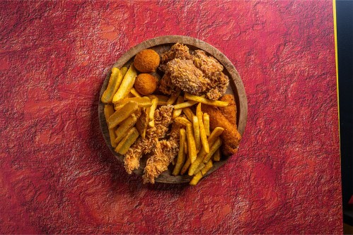 A wooden platter filled with fried chicken, golden fries, and crispy croquettes on a textured red background.