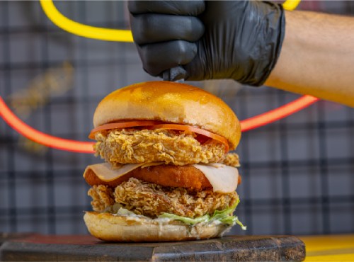 A hand in a black glove presses down on a stacked chicken sandwich with lettuce, tomato, and crispy chicken layers.