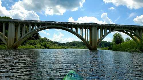 A kayak view of a large arch bridge over a calm river, surrounded by lush greenery and a blue sky with clouds.