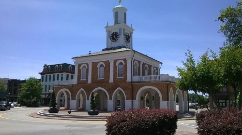 Historic building with a clock tower, arched entryways, and surrounding greenery on a sunny day.