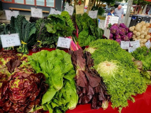 A vibrant display of various fresh greens and vegetables at a market, including kale, lettuce, and purple cabbage.