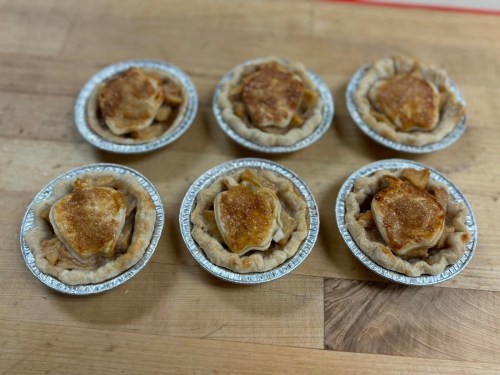 Six mini apple pies in aluminum tins, each topped with a golden-brown crust and a sprinkle of sugar.