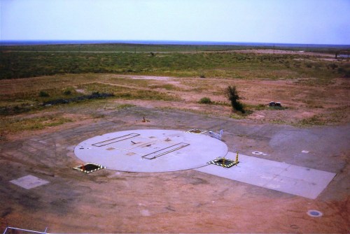 A large, empty concrete landing pad in a vast, arid landscape with sparse vegetation and distant horizons.