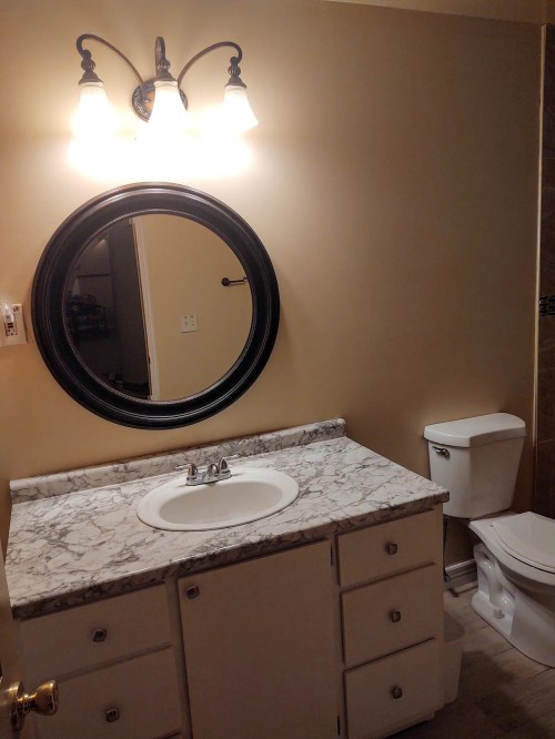 A bathroom with a marble countertop, round mirror, light fixture, and a toilet in a neutral-colored room.