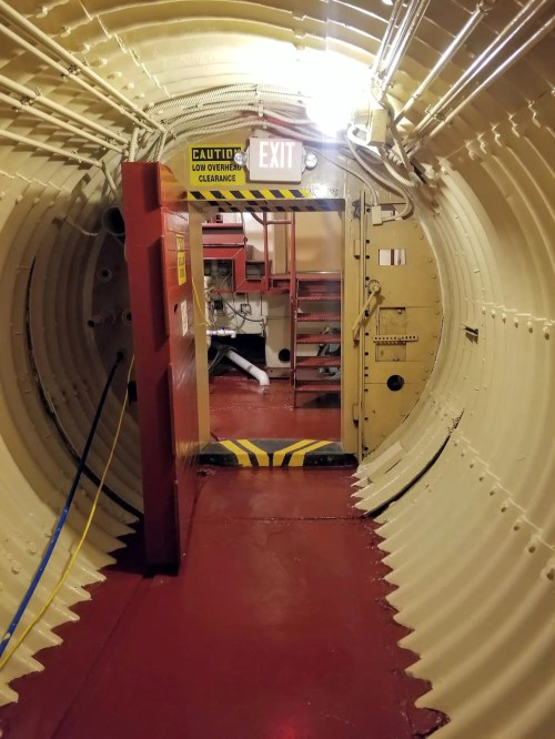 A narrow, circular hallway with a red floor, leading to an exit door marked "CAUTION" and various equipment in the background.