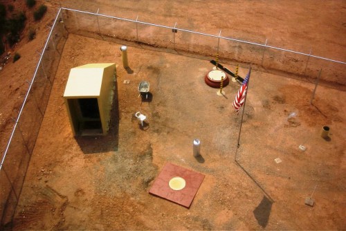Aerial view of a fenced area with a small building, flagpole, and various equipment on a dirt surface.
