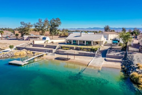 Aerial view of a waterfront home with a dock, surrounded by greenery and mountains in the background under a clear blue sky.