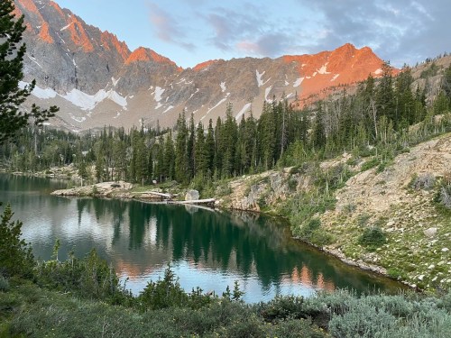 A serene lake surrounded by lush trees and mountains, reflecting the colorful sky at sunset.