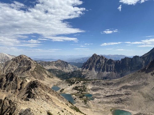 A panoramic view of rugged mountains and valleys, with clear blue lakes and a bright sky dotted with clouds.