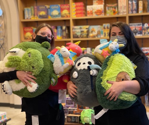 Two people hold large, colorful plush toys in a store filled with games and toys.