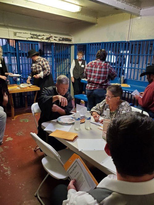 A group of people in a jail setting, engaged in discussions and activities at tables with papers and water bottles.