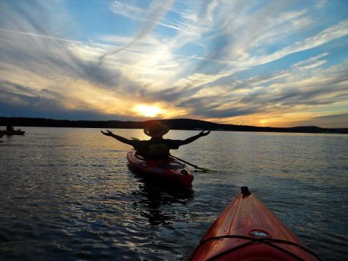 A person in a kayak with arms outstretched, silhouetted against a colorful sunset over calm water.
