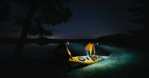 Two people in yellow jackets prepare a kayak by a calm lake under a starry night sky, using headlamps for light.