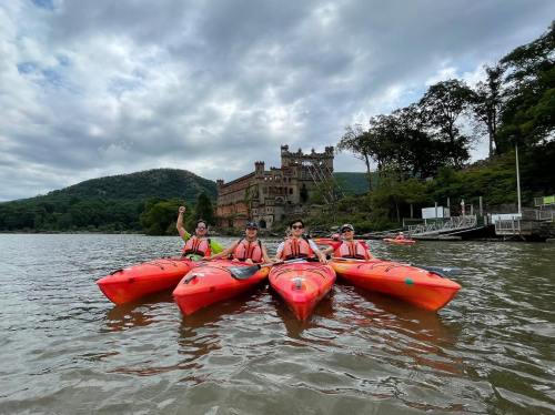 Four kayakers in bright orange kayaks on a lake, with a castle-like structure and hills in the background under a cloudy sky.