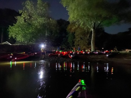 A nighttime scene of kayaks on a calm water surface, illuminated by lights, with trees and people in the background.