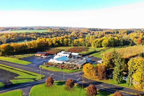 Aerial view of a farm with greenhouses, surrounded by colorful autumn trees and fields. Roads intersect nearby.