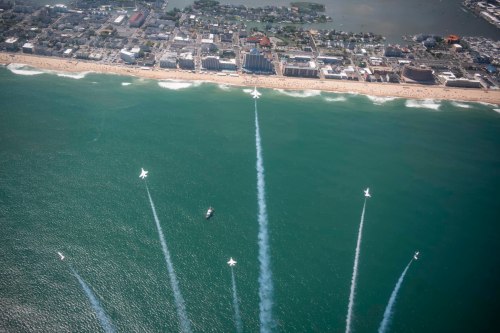 Aerial view of a beach with a city skyline, featuring planes leaving smoke trails over the ocean.
