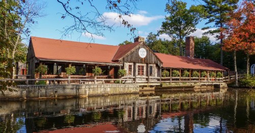 A rustic wooden building with a red roof, reflecting in a calm lake, surrounded by trees and blue skies.