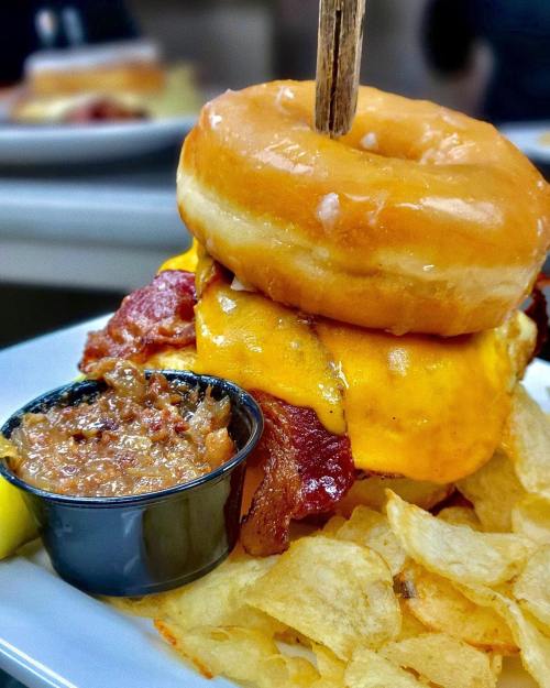A towering burger with melted cheese, bacon, and a glazed donut on top, served with a side of chips and sauce.