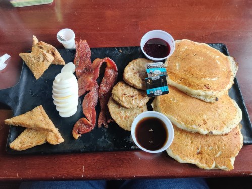 A breakfast platter featuring pancakes, bacon, sausage, toast, and syrup served on a black plate.