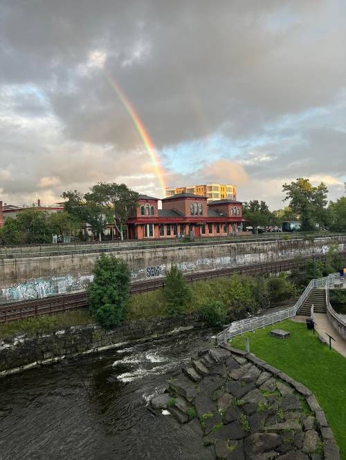 A rainbow arcs over a riverside building, with greenery and a rocky bank visible along the water's edge.