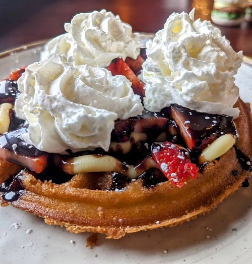 A golden waffle topped with whipped cream, strawberries, chocolate sauce, and banana slices on a white plate.