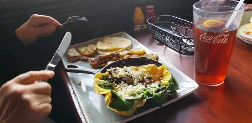 A plate with a tortilla filled with greens and meat, alongside potatoes and a drink, on a wooden table.