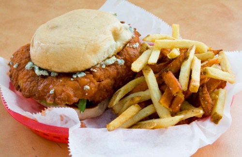 A crispy fried chicken sandwich with blue cheese on a bun, served with a side of golden fries in a red basket.