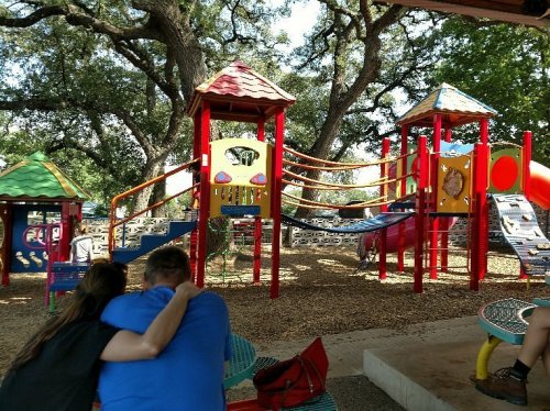 A couple sits together at a playground with colorful play structures and trees in the background.