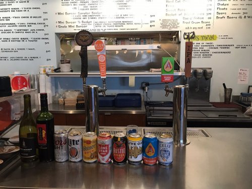 A bar counter displaying various drinks, including wine, beer cans, and two beer taps, with a menu in the background.