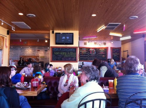 A busy restaurant interior with patrons seated at tables, colorful decor, and a menu board in the background.
