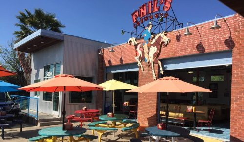 Colorful outdoor seating area at Phil's House, featuring umbrellas and a cowboy-themed sign. Bright, sunny day.