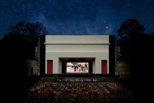 An outdoor theater at night, featuring a large screen and an audience seated in front, under a starry sky.