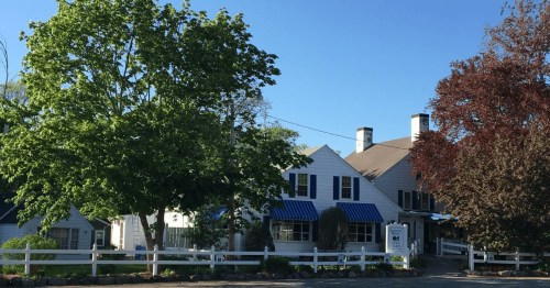 A charming white house with blue awnings, surrounded by trees and a white fence, under a clear blue sky.