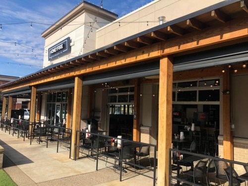 Exterior view of a restaurant with wooden beams, outdoor seating, and large open windows under a blue sky.