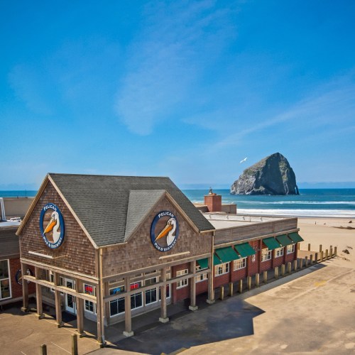 A beachside restaurant with a logo, overlooking a sandy shore and a large rock formation in the distance under a clear blue sky.