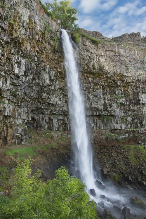 A tall waterfall cascades down a rocky cliff, surrounded by lush greenery and a blue sky with scattered clouds.