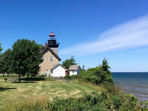 A lighthouse stands near the shore, surrounded by green trees and a clear blue sky over the calm water.