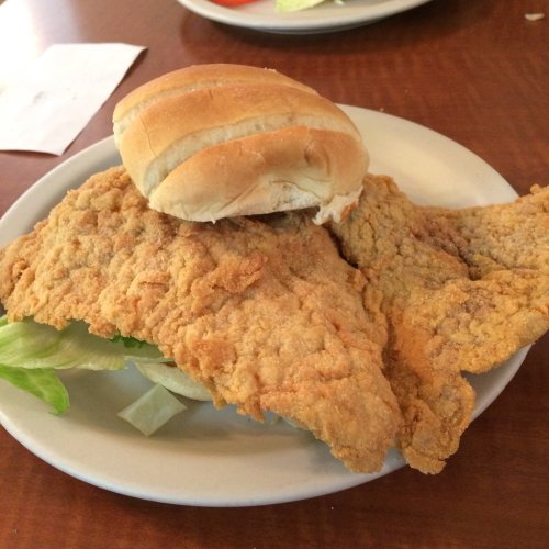 A plate with two pieces of fried fish, lettuce, and a soft bread roll on a wooden table.