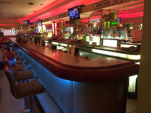 A brightly lit bar with a long counter, stools, and colorful neon signs in a lively atmosphere.