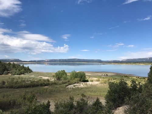 A serene lake surrounded by greenery and distant mountains under a blue sky with scattered clouds.