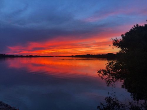 A serene lake at sunset, reflecting vibrant orange and purple hues in the sky and water, surrounded by trees.
