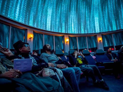 A diverse group of people sits in a planetarium, gazing up at a vibrant, illuminated dome display.