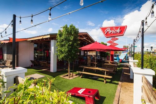 Outdoor dining area of The Porch restaurant, featuring picnic tables, umbrellas, and string lights under a blue sky.