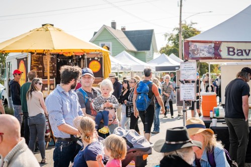 A busy outdoor market scene with people browsing stalls, food trucks, and families enjoying the day.